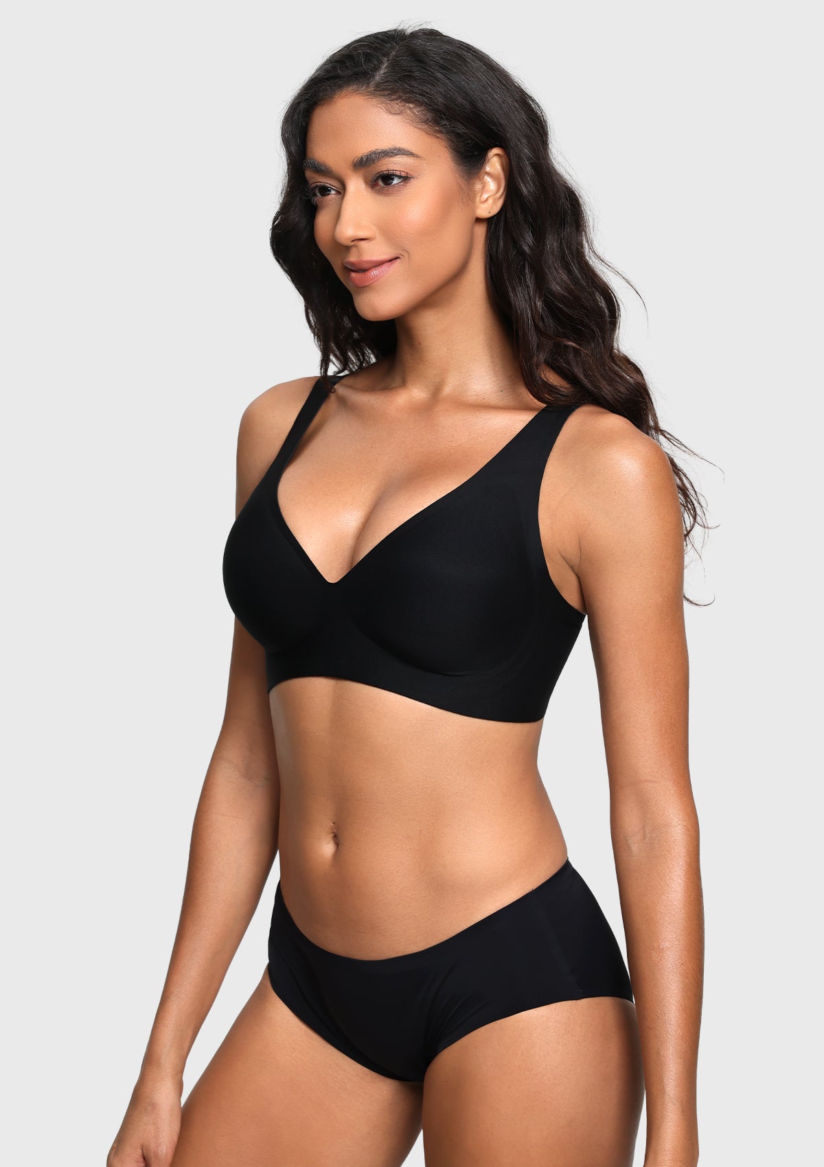 A woman modeling a black two-piece lingerie set consisting of a bra and matching underwear, photographed against a solid light gray background.