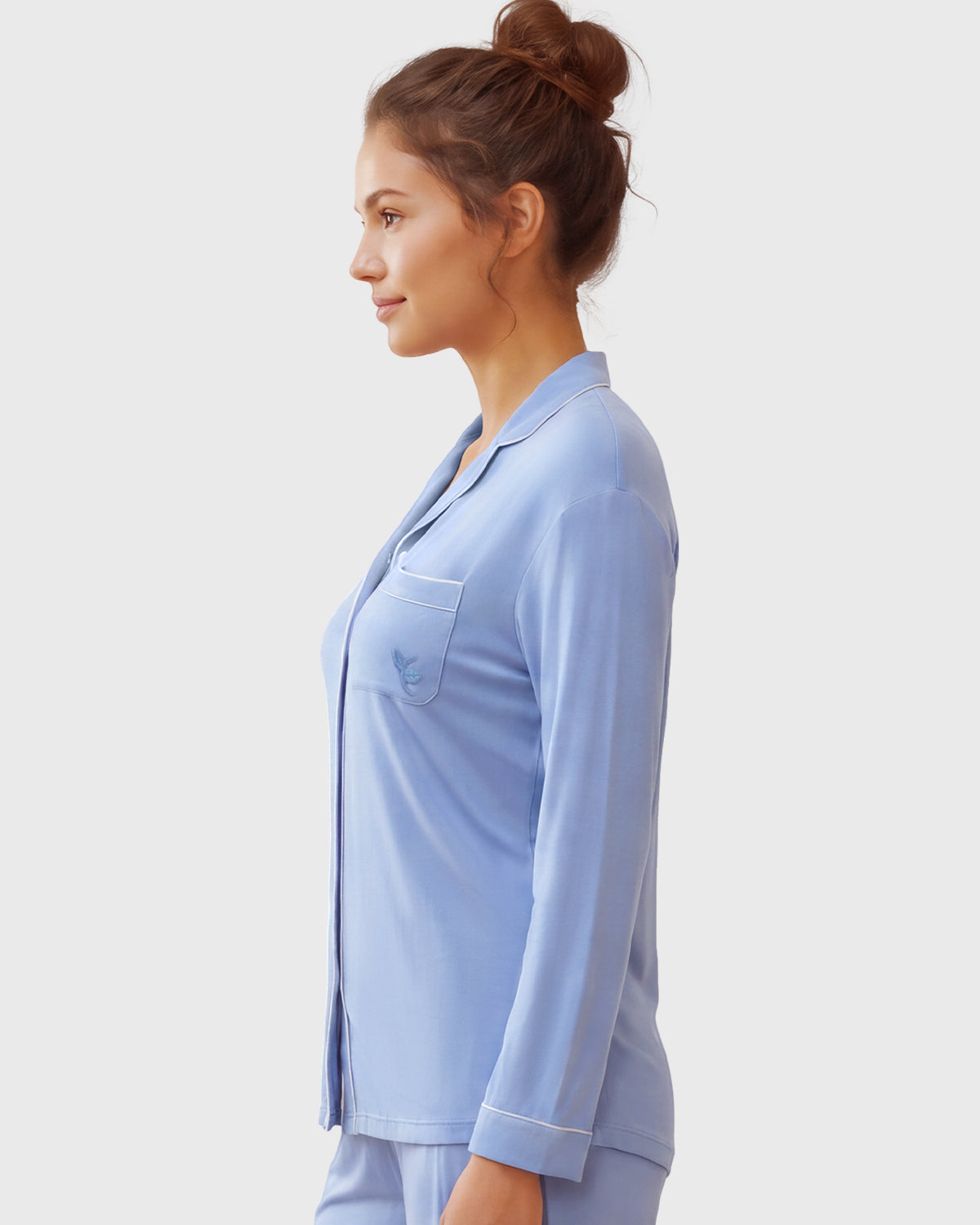 A woman in profile wearing a light blue pajama set with a long-sleeve button-front top and matching pants, photographed against a plain light gray studio background.