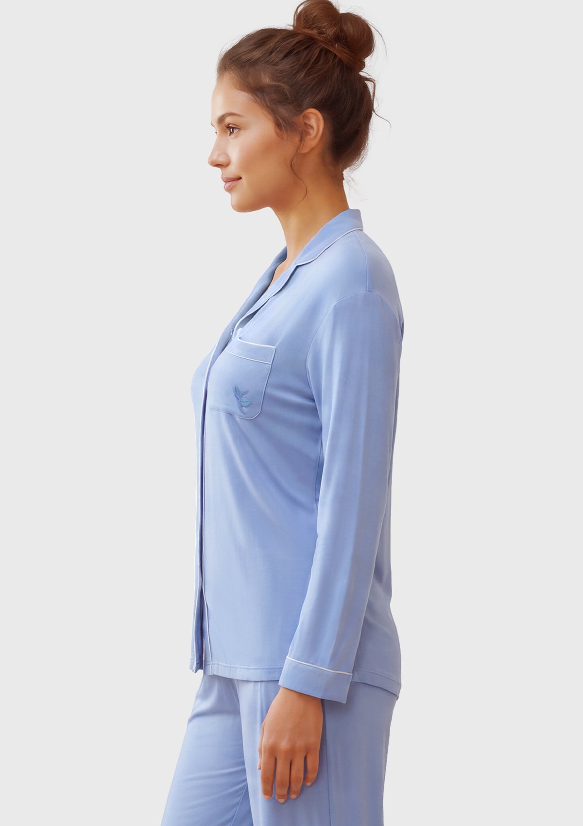 A woman in profile wearing a light blue pajama set with a long-sleeve button-front top and matching pants, photographed against a plain light gray studio background.