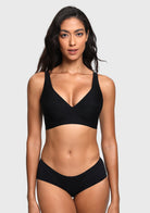 A woman modeling a black two-piece bikini set against a light gray studio background. The set includes a V-neck bikini top with wide shoulder straps and matching high-cut bikini bottoms.