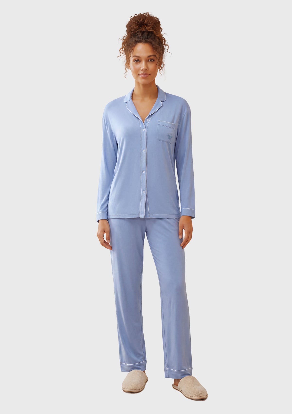 A woman modeling a light blue long-sleeve pajama set with a button-down shirt and matching pants, paired with beige slippers, against a light gray studio background.