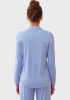 A woman viewed from the back wearing a light blue long-sleeve collared top and matching light blue pants against a plain light gray studio background.