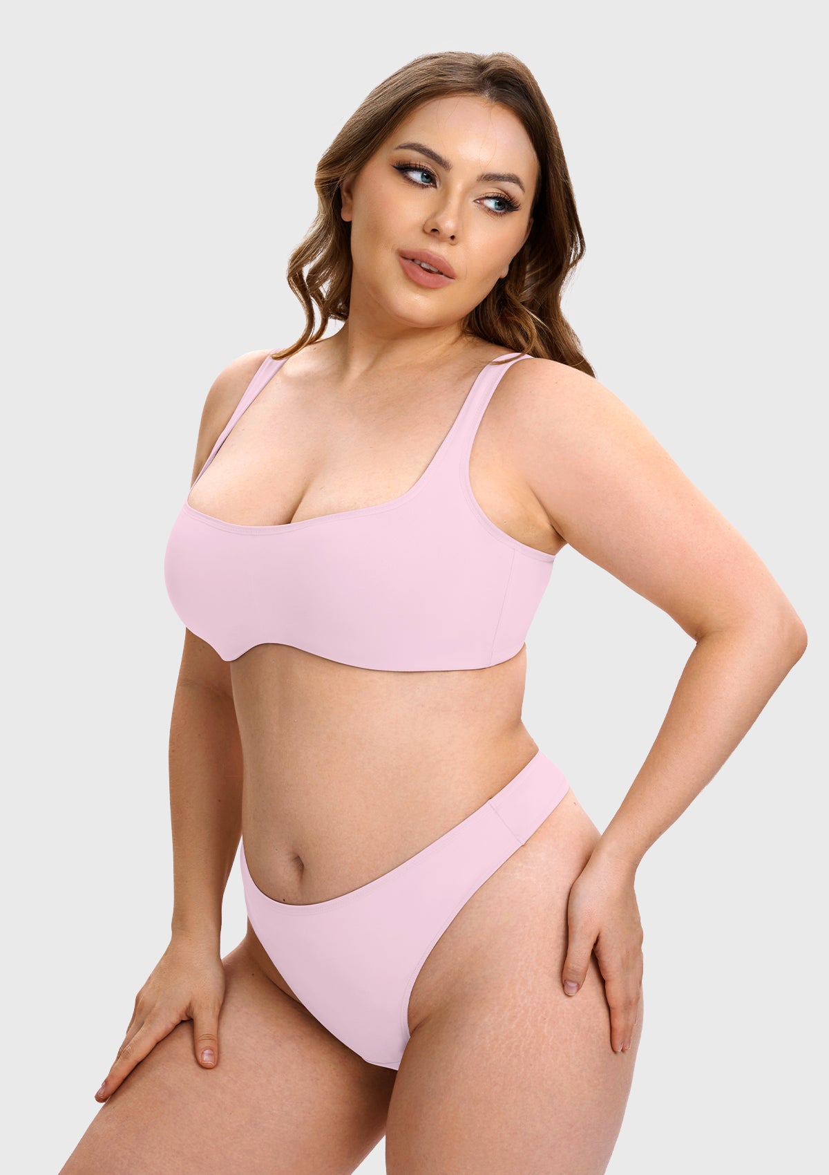 A plus-size model wearing a light pink two-piece bikini set, consisting of a scoop-neck bralette and matching high-cut bottoms, against a solid light gray background.