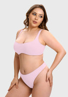 A plus-size model wearing a light pink two-piece bikini set, consisting of a scoop-neck bralette and matching high-cut bottoms, against a solid light gray background.