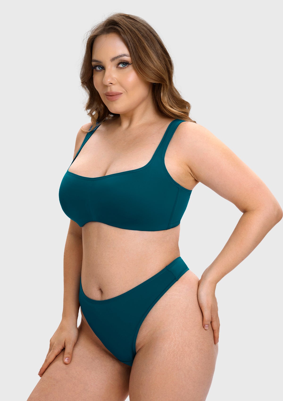 A woman modeling a two-piece high-cut bikini set in a deep teal color against a solid light gray background.