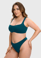 A woman modeling a two-piece high-cut bikini set in a deep teal color against a solid light gray background.