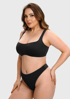 A woman modeling a black two-piece bikini set against a light gray studio background. The set includes a scoop-neck bra top and matching high-cut bottoms.