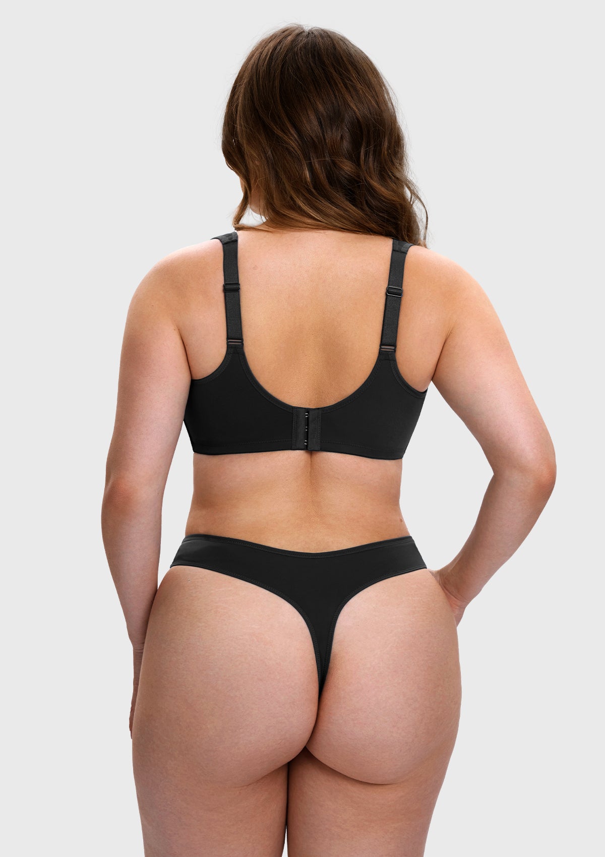 A woman shown from the back wearing a black bra and matching thong underwear against a light gray studio background.