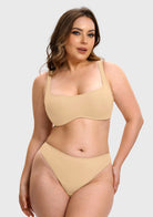 A woman modeling a two-piece beige underwear set consisting of a scoop-neck bra and matching high-cut briefs, set against a solid light gray background.